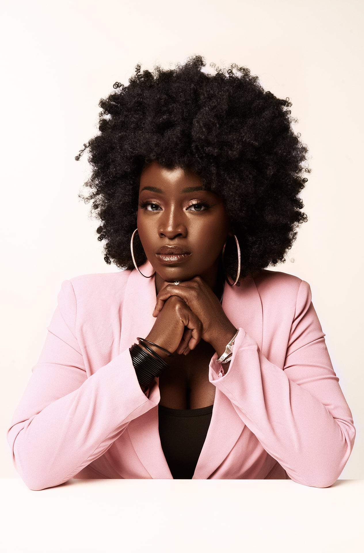 Portrait of Lovette Jallow, a Black woman with medium length hair, impeccable skin, and wearing a pink blazer and hoops.